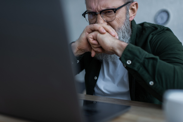 homme âgé stressé dans les lunettes fronçant les sourcils et regardant flou ordinateur portable à la maison - Photo, image