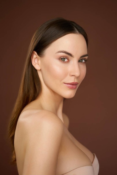 Vista lateral de atraente jovem modelo feminino com ombros nus e cabelos castanhos olhando para a câmera contra fundo marrom - Foto, Imagem