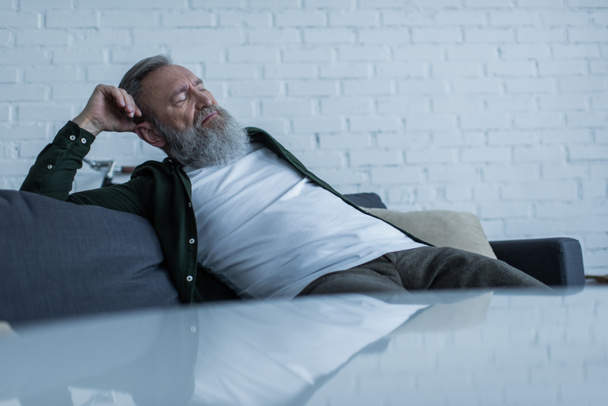 järkyttynyt vanhempi mies parta kärsii kriisistä istuessaan sohvalla olohuoneessa  - Valokuva, kuva