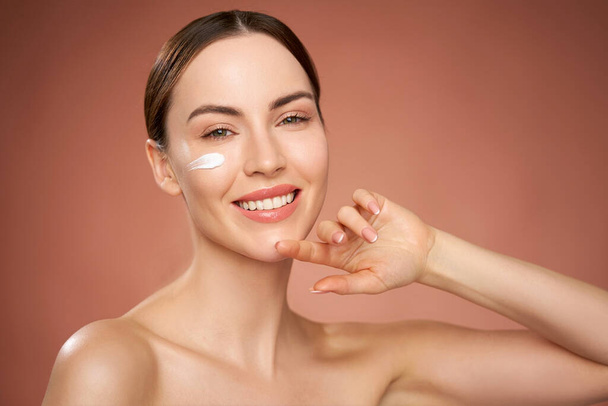 Happy young female with moisture cream applied under eye smiling and looking at camera during skin care routine against brown background - Photo, Image