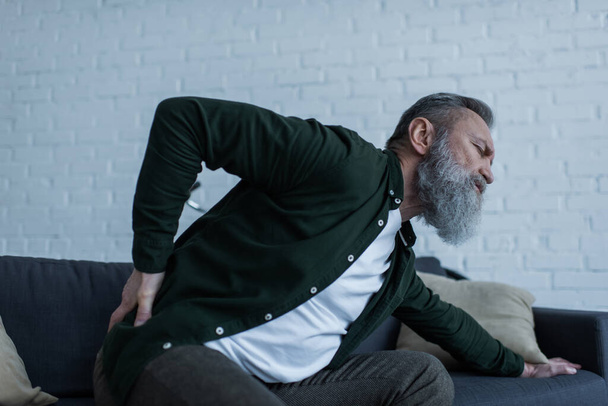 bearded senior man with beard sitting on sofa and touching back while suffering from pain  - Photo, Image