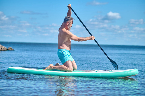 Summer pastime. Adult grey-haired bearded man kneeling sideways to camera paddling on board on water on sunny day - Valokuva, kuva
