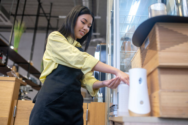 Higiene en el trabajo. Vista inferior de la joven mujer asiática en delantal tocando desinfectante sonriendo de pie en el lugar de trabajo en la cafetería - Foto, Imagen