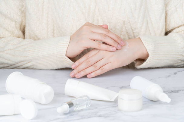 Γυναίκα εφαρμογή ενυδατική κρέμα στα χέρια της με προϊόντα περιποίησης του δέρματος σε μαρμάρινο τραπέζι. Έννοια ρουτίνας φροντίδας του δέρματος. - Φωτογραφία, εικόνα