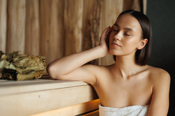 Woman with closed eyes sitting in wooden sauna - Valokuva, kuva