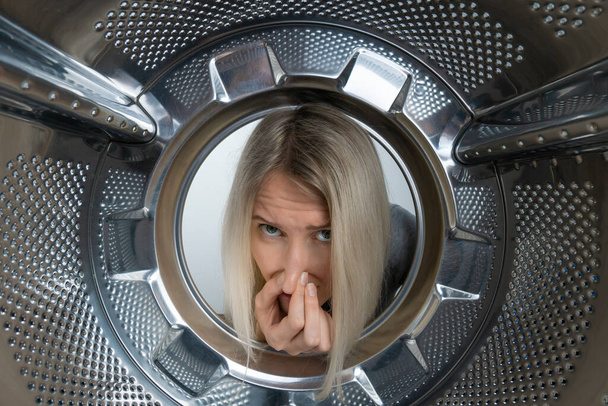 Young blonde woman looks into the drum of a washing machine and holds her nose against an unpleasant smell, photo from the inside - Фото, изображение