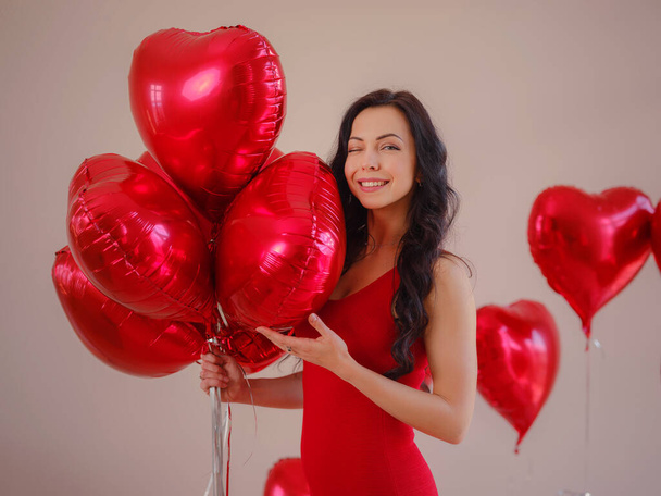 Jovem feliz bonita em vestido vermelho. Festa de férias. Modelo alegre posando com balões em forma de coração vermelho, se divertindo, comemorando o Dia dos Namorados. Símbolo do amor - Foto, Imagem