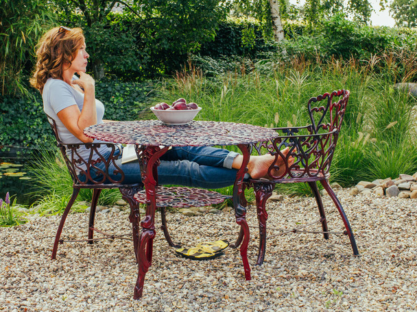 Een volwassen vrouw met een kom vol verse pruimen staat op een metalen tafel in een buitentuin, op haar eigen grondgebied, ontspannen in haar vrije tijd. - Foto, afbeelding