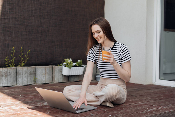 Smiling young woman sitting outdoor on terrace, working remotely online on laptop and drink fresh juice side view. Freelance, distant job, business lady. Working from home in summer time. - Photo, Image