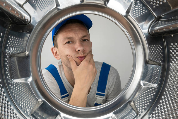 Male repairman in uniform holds his chin and looks thoughtfully into the camera, photo from inside the drum of a washing machine - Foto, Imagem