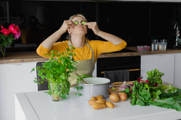エプロンの面白いブロンドの女性は台所でマスクとして目にキュウリのスライスを保持します。スープやサラダ用の新鮮な野菜。ジャガイモ、緑、テーブルの上のパンの近くのビートルート。家庭での美容治療 - 写真・画像