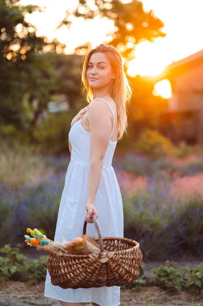 Young Woman in White Dress and Basket in Her Hands stands in Lavender Field at Sunset. Vertical Portrait of Girl at Golden Hour - Fotografie, Obrázek