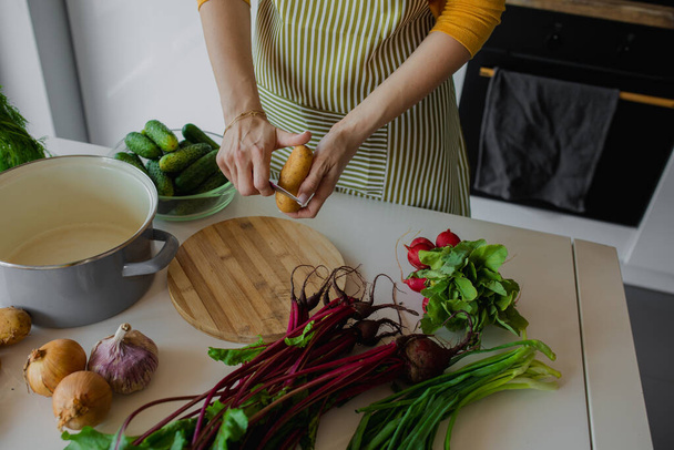 Cropped woman in apron peeling potato and chop fresh vegetables on wooden board in kitchen. Cooking mixed summer salad or soup. Cucumber, onion, beetroot near pan as lunch on table. Raw food recipe - Photo, image