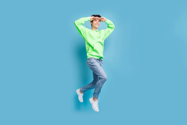 Photo of attractive curious gentleman wear green sweatshirt jumping looking far away empty space isolated blue color background. - 写真・画像