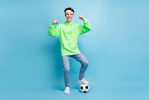 Foto van koele zelfverzekerde jonge man gekleed groene hoodie spelen voetbal tonen kracht glimlachen geïsoleerde blauwe kleur achtergrond. - Foto, afbeelding