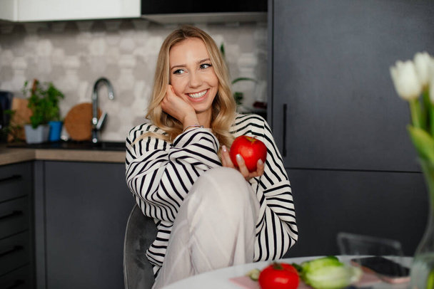 Cute Blonde lady Holding apple in hand. Young european woman at kitchen indoors with healthy food. Dieting concept - Фото, изображение