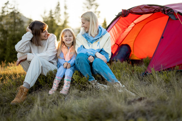 Duas irmãs ou amigos se sentam junto com uma menina e conversam no acampamento, sentados felizes perto da tenda ao pôr-do-sol. Família homossexual com uma criança viajar nas montanhas - Foto, Imagem