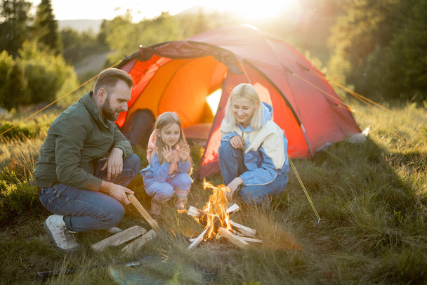 Young adult couple sit with a little girl by the campfire at campsite, traveling with tent on nature. Family spend summer time hiking in the mountains - Photo, Image