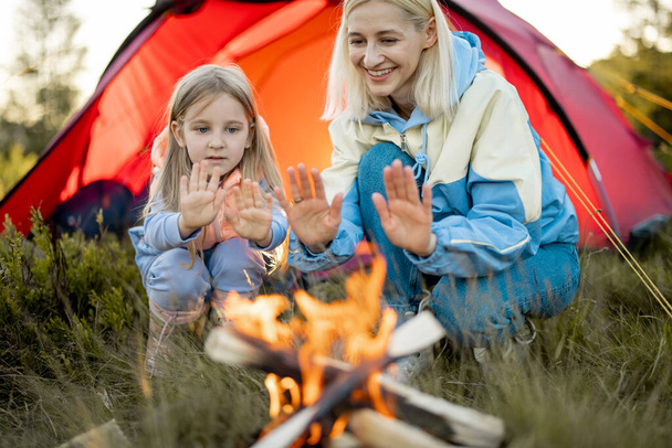 Young woman with her little girl sit by the campfire, bonding together while travel on nature. Mother with daughter warming up at campsite - Photo, Image