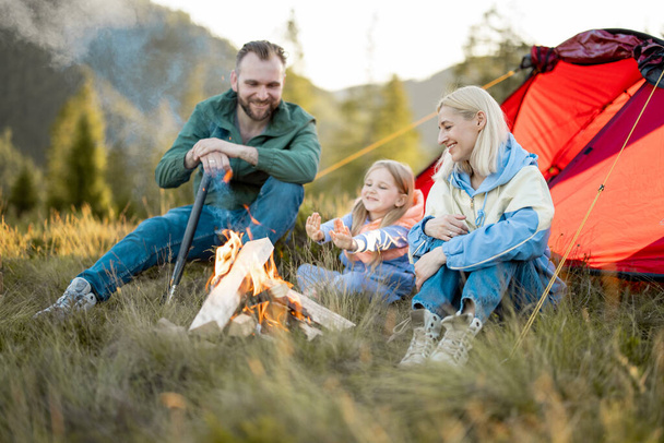 Young adult couple sit with a little girl and have lovely conversation by the campfire, traveling with tent on nature. Family spend summer time hiking in the mountains - Foto, Imagem