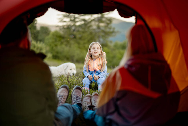 Schattig klein meisje vertellen een verhaal aan haar ouders die zitten in campingtent, Uitzicht van de binnenkant van de tent. Jeugd, verhalen vertellen en verbondenheid met het natuurconcept - Foto, afbeelding