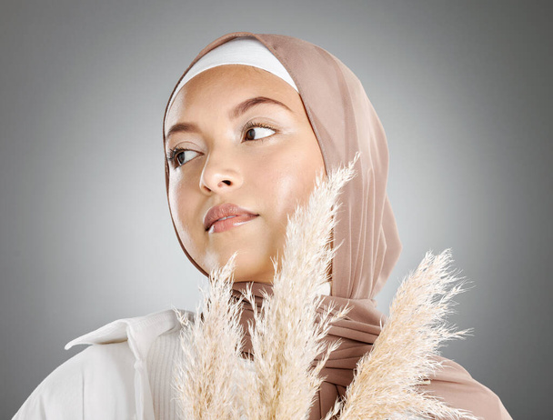Une belle jeune femme musulmane portant un foulard brun tenant une plante de blé pampas isolée sur fond de studio gris. Modeste arabe féminin maquillé avec le visage recouvert de hijab traditionnel. - Photo, image