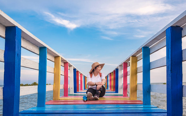 Перспективний вигляд щасливої жінки-туристки, дивлячись збоку, сидячи на барвистому дерев'яному мосту на морському тлі хмари на фоні блакитного неба
 - Фото, зображення