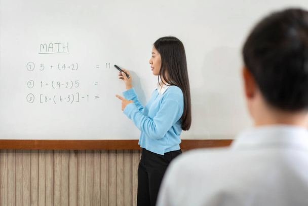 Női oktató áll a tábla előtt, és matematikai egyenleteket ír a fedélzetre, hogy elmagyarázza a diáknak matekórán az osztályteremben. - Fotó, kép