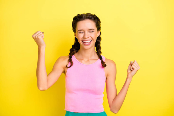 Photo of shiny lucky young lady braids dressed singlet rising fists closed eyes isolated yellow color background. - Foto, Imagen