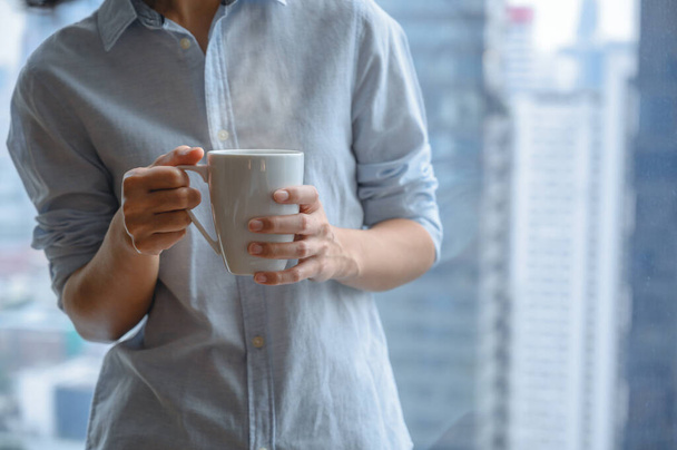 Primo piano mani di donna in possesso di una tazza di caffè o tè bianco dalla finestra con sfocato di vista sulla città alto edificio sullo sfondo. Copia spazio - Foto, immagini