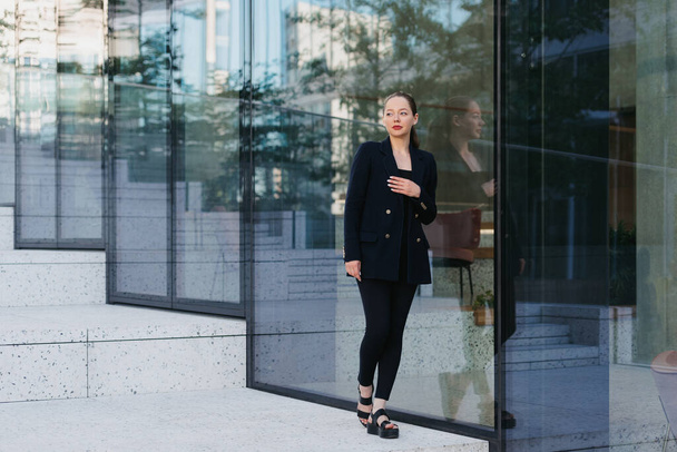Una foto completa de una señora en una chaqueta que está apoyada contra la pared de cristal en el distrito financiero. Una mujer de negocios con el brazo en el pecho en el espacio urbano contemporáneo. - Foto, Imagen