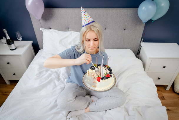 Sad, upset woman in pajama and party cap eating by spoon her birthday cake sitting on the bed in decorated bedroom. Celebrates birthday alone. Jamming of negative emotions. Selective focus - Fotoğraf, Görsel