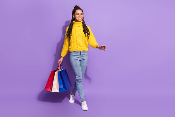 Full size photo of optimistic brunette lady hold bags go wear yellow sweater jeans sneakers isolated on purple color background. - Foto, Imagen