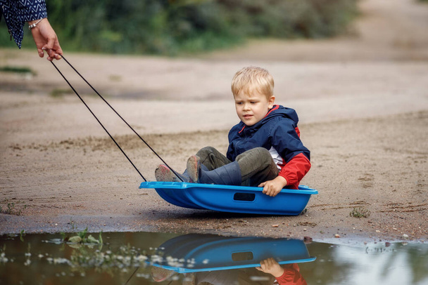 Mały chłopiec siedzący na sankach w błocie koło bagien. Lato po deszczu, sanki holowania jego mama żartują. - Zdjęcie, obraz
