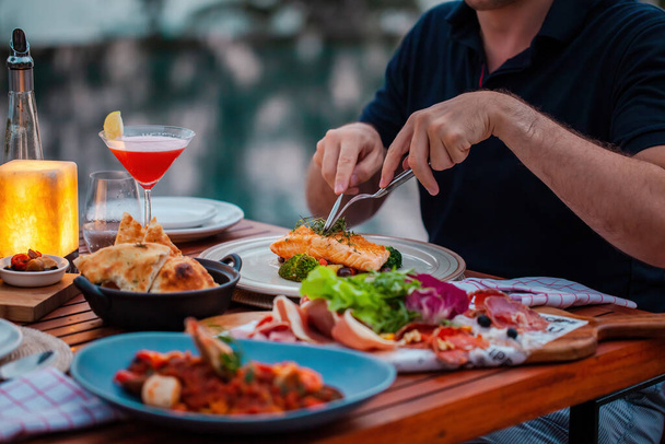 Мужчина на роскошный ужин подается на столе со свежими морепродуктами, холодными нарезками и коктейльным бокалом в роскошном открытом ресторане. Мужские руки режут лосося на гриле. Здоровое питание - Фото, изображение