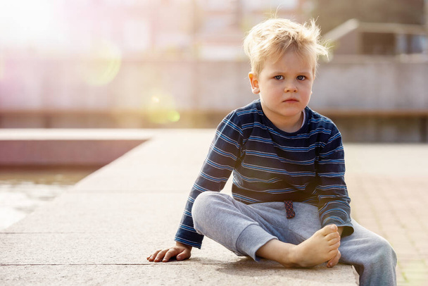 Маленький грустный мальчик сидит у фонтана в солнечный день, ребенок смотрит в фотокамеру, на фотографии есть свободное место для текста. Летний отдых для детей в городе. - Фото, изображение