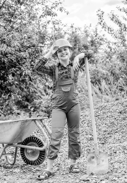 cheerful child laborer using building uniform and shovel and construction wheelbarrow, childhood. - Foto, afbeelding