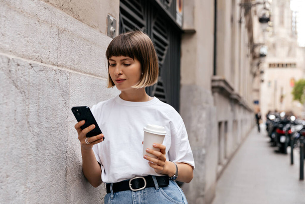 Niesamowita urocza kobieta z krótkimi włosami, nosząca biały t-shirt przewijający smartfon w mieście. Młoda urocza dama z kawą i telefonem w mieście.  - Zdjęcie, obraz