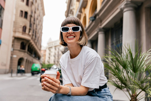 Riendo encantadora dama con pelo corto con gafas de sol blancas en camiseta blanca sosteniendo café y posando al aire libre en un día soleado y cálido en la ciudad europea.  - Foto, Imagen
