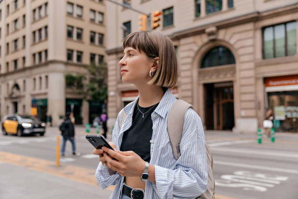 Spectacular happy caucasian girl with short hair wearing blue shirt with backpack is holding smartphone and looking aside on city background in warm day.  - Photo, Image