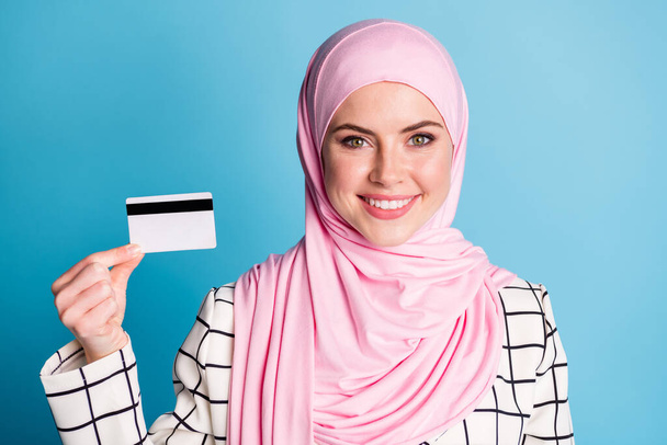 Close-up portrait of pretty content cheerful muslimah girl holding in hand bank card finance isolated over bright blue color background. - Fotoğraf, Görsel