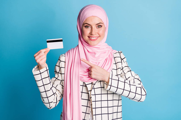 Πορτρέτο του περιεχομένου χαρούμενα muslimah κορίτσι κρατώντας στο χέρι αποδεικνύοντας Cashback τραπεζική κάρτα απομονώνονται σε φωτεινό μπλε χρώμα φόντο. - Φωτογραφία, εικόνα