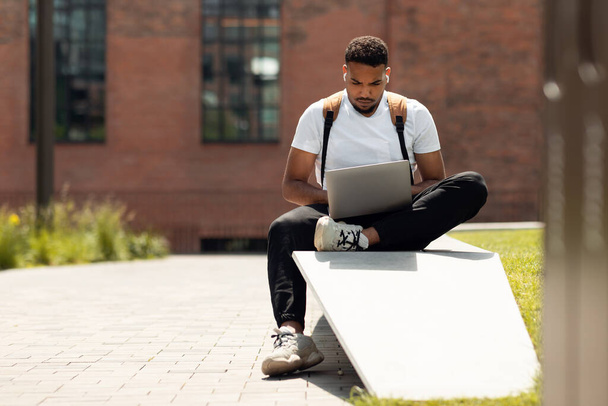 Ελεύθερος επαγγελματίας. Trendy Αφρικής Αμερικανός άνθρωπος χρησιμοποιώντας φορητό υπολογιστή σε εξωτερικούς χώρους, που εργάζονται σε απευθείας σύνδεση, ενώ κάθεται στον πάγκο στην αστική περιοχή της πόλης, ελεύθερο χώρο - Φωτογραφία, εικόνα