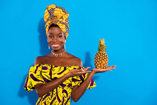 Photo of young sweet cheerful african woman in turban recommending pineapple isolated on blue color background. - Zdjęcie, obraz