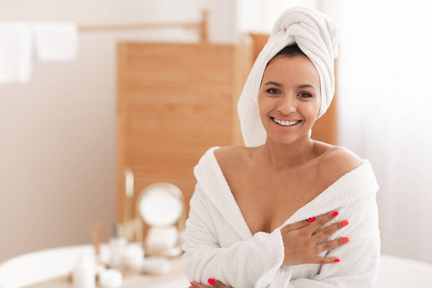 Великолепная женщина позирует с обернутым полотенцем на голове в халате, улыбаясь в камеру, стоящую в современной ванной комнате в помещении. Концепция косметики и ухода за телом - Фото, изображение