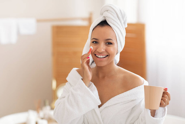 Skincare. Happy Lady Applying Facial Moisturizer On Cheek And Holding Coffee Mug Smiling To Camera Posing In Modern Bathroom At Home. Morning Beauty Care Routine Concept - Фото, зображення