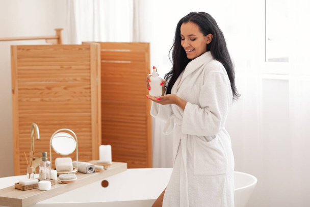 Cosméticos para el cuidado corporal. Mujer alegre usando loción corporal o jabón líquido cuidándose a sí misma disfrutando de la rutina de belleza de pie en el baño moderno en casa. Bienestar y Spa - Foto, Imagen