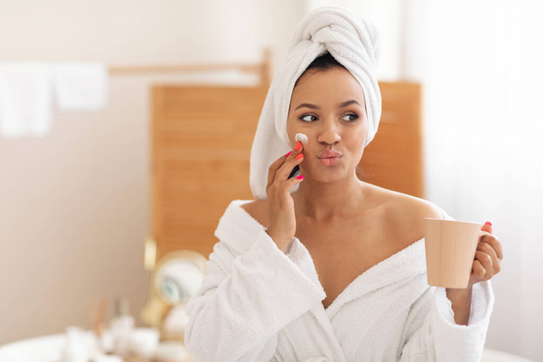 Pielęgnacja twarzy. Zabawna kobieta stosująca krem nawilżający na twarzy wylewanie warg pozowanie z owiniętym ręcznikiem na głowie trzymając filiżankę kawy w łazience w domu rano. Pielęgnacja skóry Beauty produktu - Zdjęcie, obraz