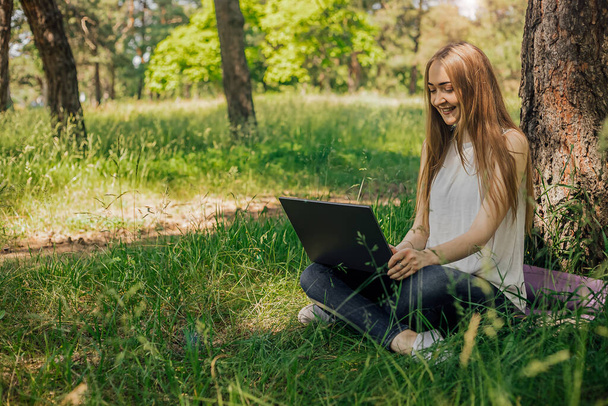 En la pancarta, una joven trabaja con una computadora portátil al aire libre en el parque, sentada en el césped. El concepto de trabajo remoto. Trabaja como freelancer. La chica toma cursos en un ordenador portátil y sonríe - Foto, imagen