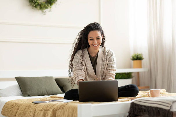Fröhliche junge Dame sitzt im Schneidersitz auf dem Bett, nutzt Laptop für Online-Arbeit oder Bildung zu Hause, Kopierraum. Schöne Millennials studieren aus der Ferne, haben Geschäftstreffen im Web - Foto, Bild